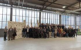 Mitarbeiter stehten in einer Werkhalle vor einer Wand mit Handabdrücken