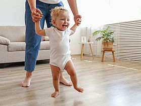 Glückliches Kleinkind macht erste Schritte mit Hilfe einer erwachsenen Person im Wohnzimmer